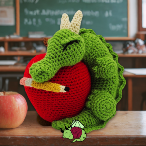 a crochet baby dragon sleeps, holding a pencil in his mouth, wrapped around an apple