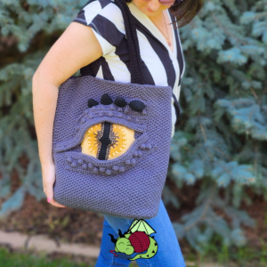 woman carrying a crochet tote bag with a large grey detailed dragon eye on one side.